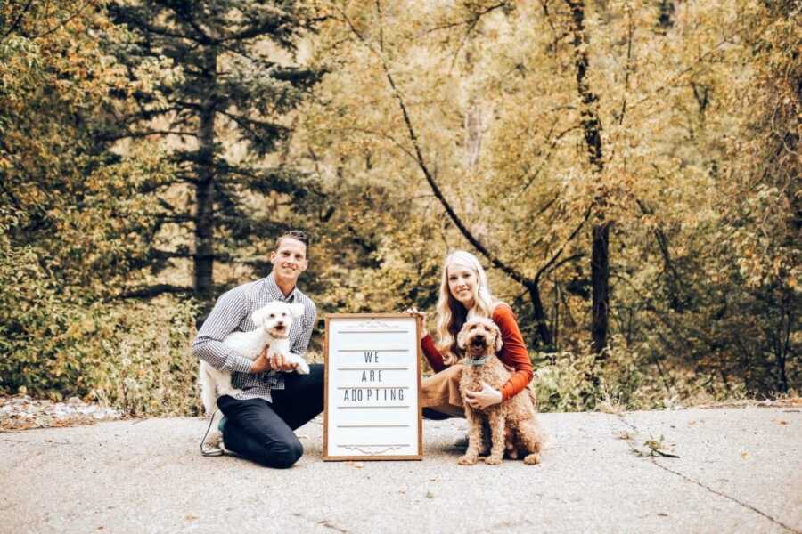 Young married couple take a photo with their two dogs while holding a sign that says "We are adopting"