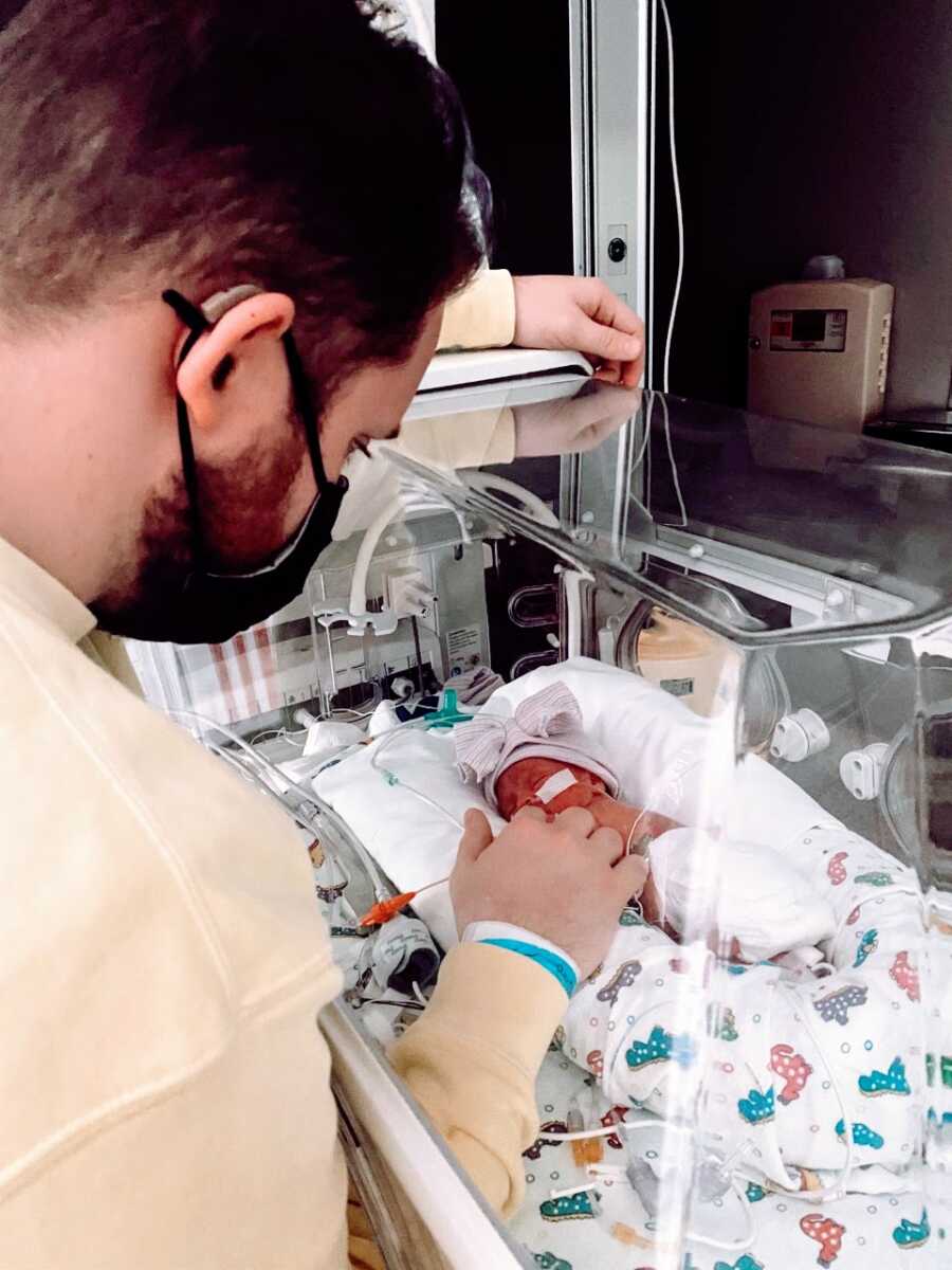 First time dad wears a face mask while watching his premature newborn daughter sleep in an isolette in the NICU