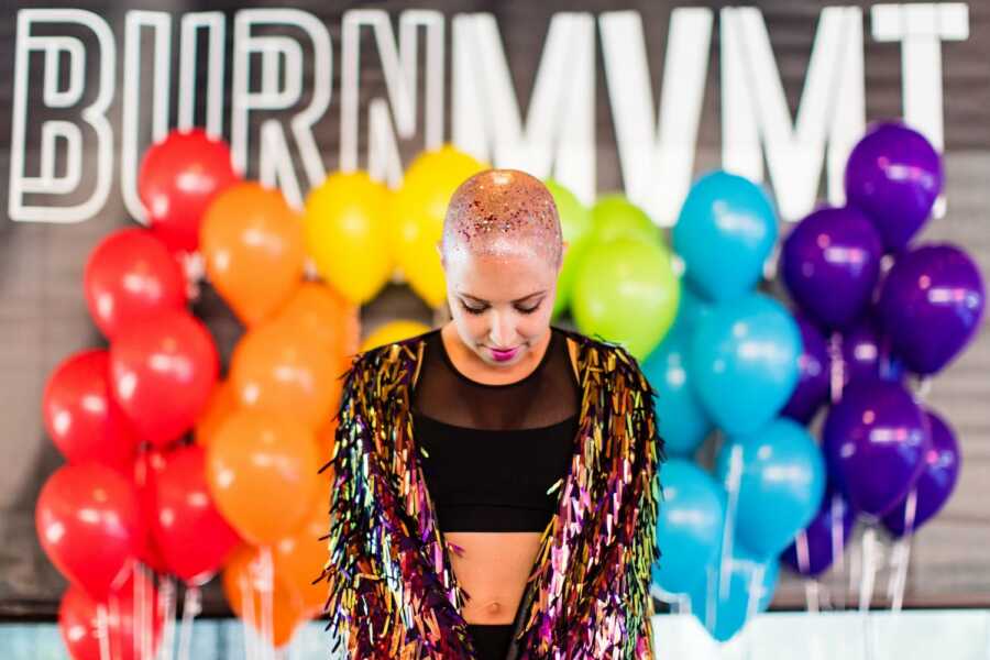 LGBT woman battling breast cancer takes a photo with glitter on her bald head and rainbow balloons behind her