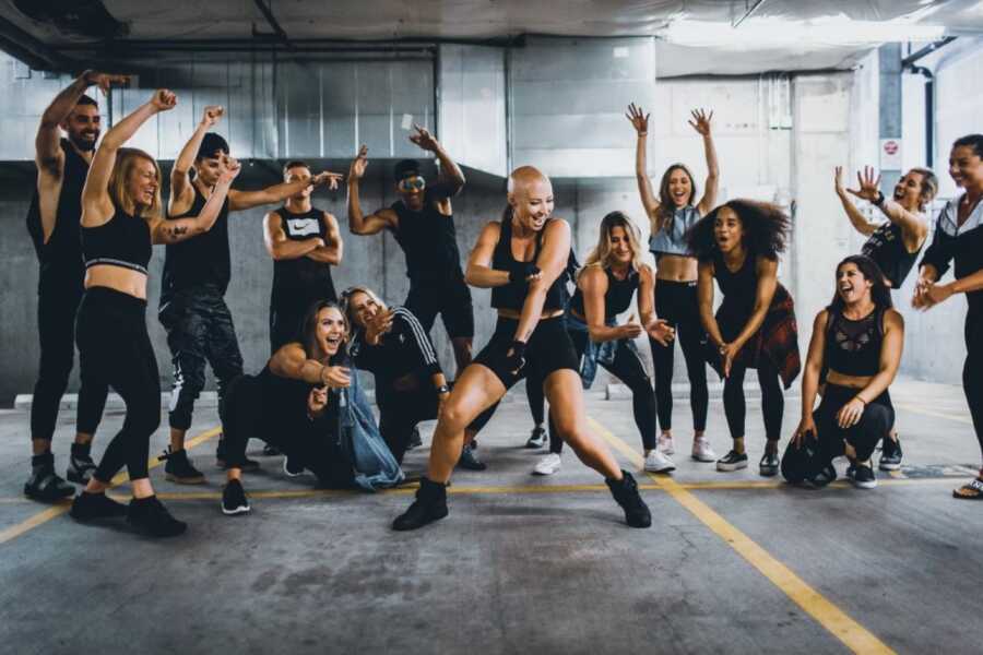 Fitness instructor battling breast cancer dances around a circle of gym-goers that she teaches