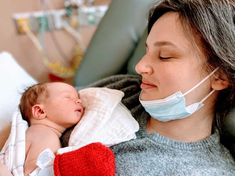 New mom wearing a gray sweater smiles down at her newborn son sleeping in her arms