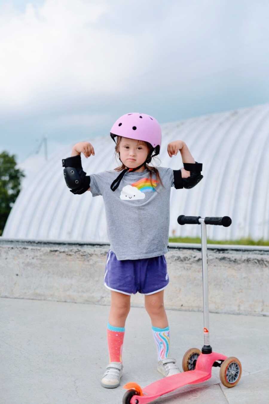 Little girl with Down Syndrome looks serious while showing off her determination and muscles to ride a scooter with a helmet and pads