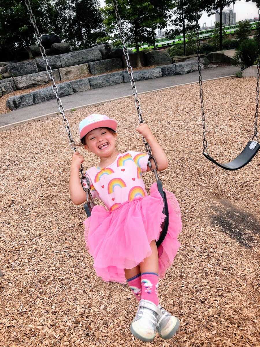 Little girl with Down Syndrome smiles big while swinging at the park in a pink ruffled dress with rainbows and hearts on it