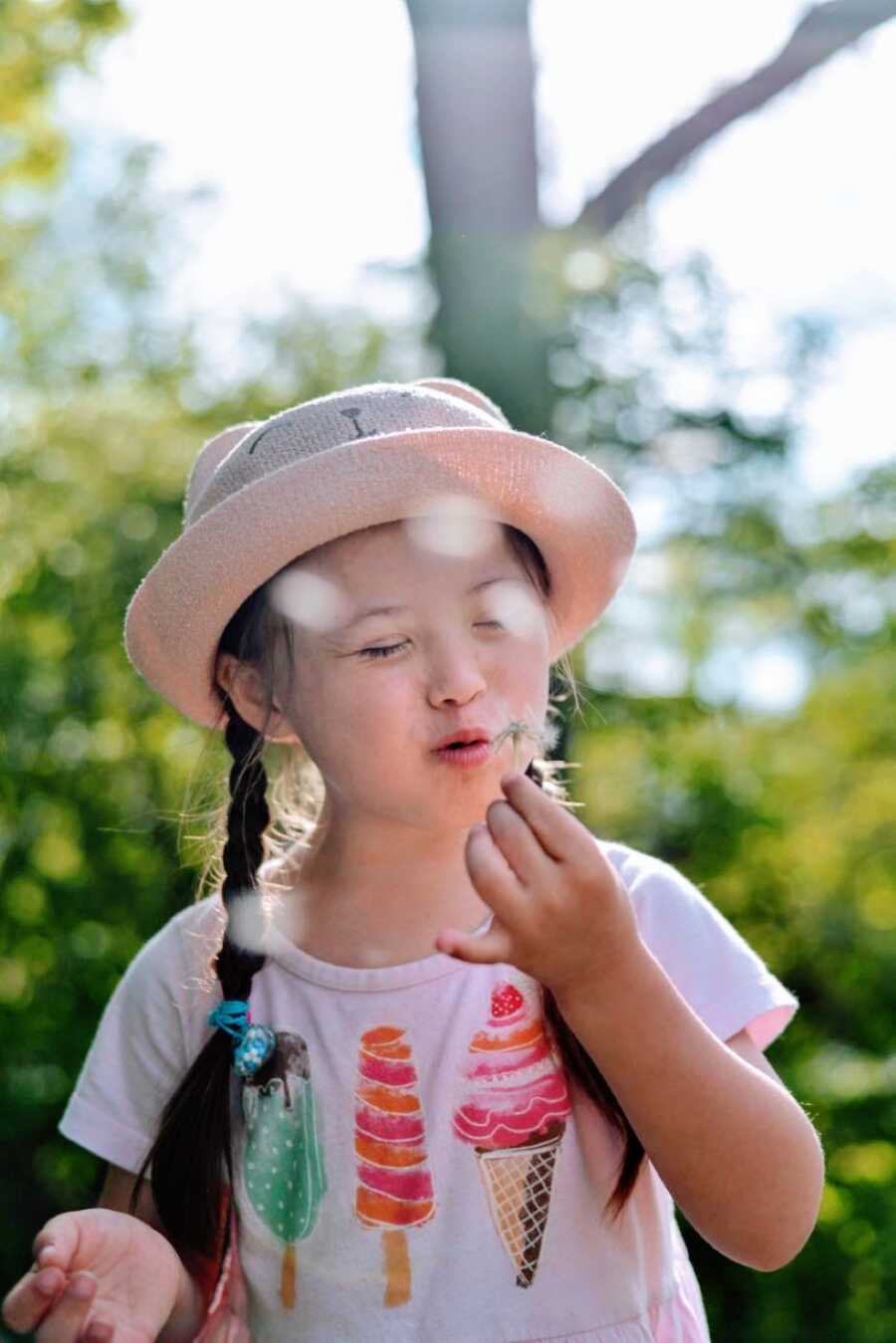 Little girl with Down Syndrome makes a wish while blowing on a dandelion in a pink shirt with a matching pink hat