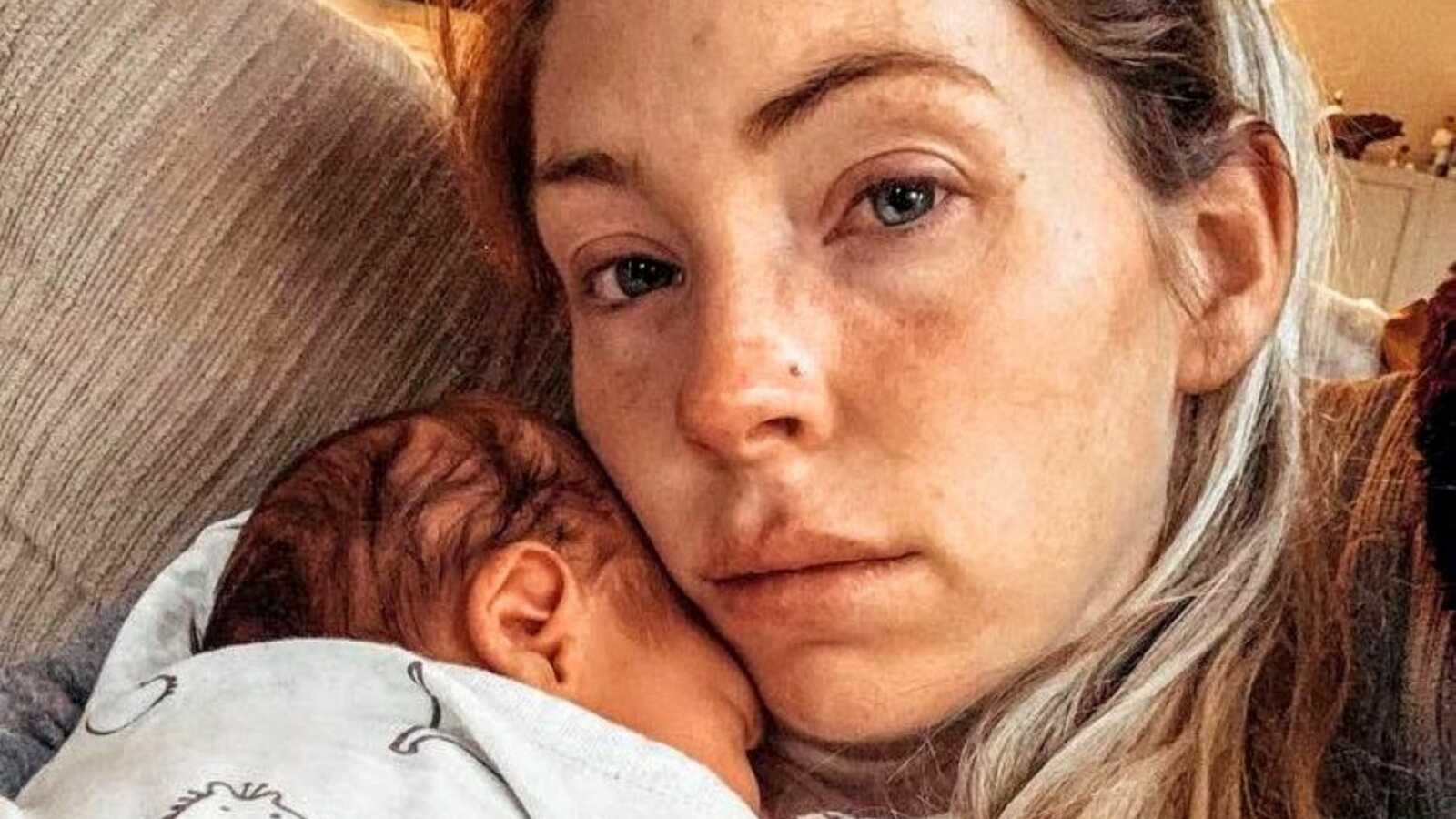 Mom suffering from postpartum anxiety holds her sleeping newborn baby on the couch while trying not to cry