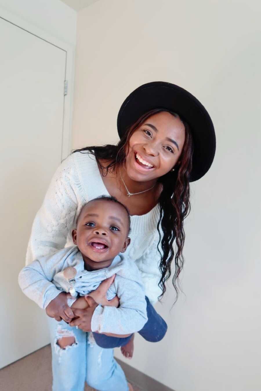 Young solo mom takes a photo with her son while she wears a white sweater and black hat and he wears a blue onesie