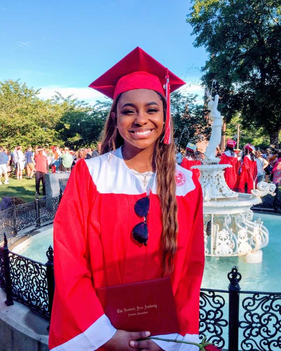 Young girl takes a photo at her high school graduation in a red cap and gown while holding her diploma