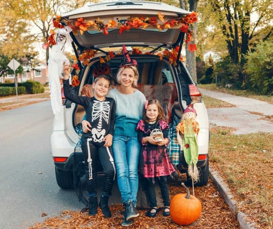 Mom and kids sit in the trunk of their car, all decorated for Halloween.