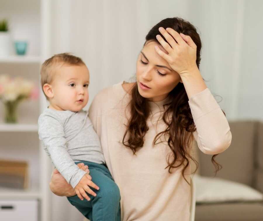 Young mother balances baby on her hip and presses her hand to her head in exhaustion.