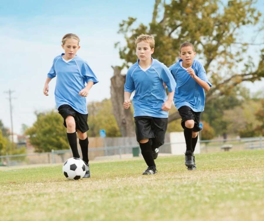 Three kids in blue jerseys chase after soccer ball. 