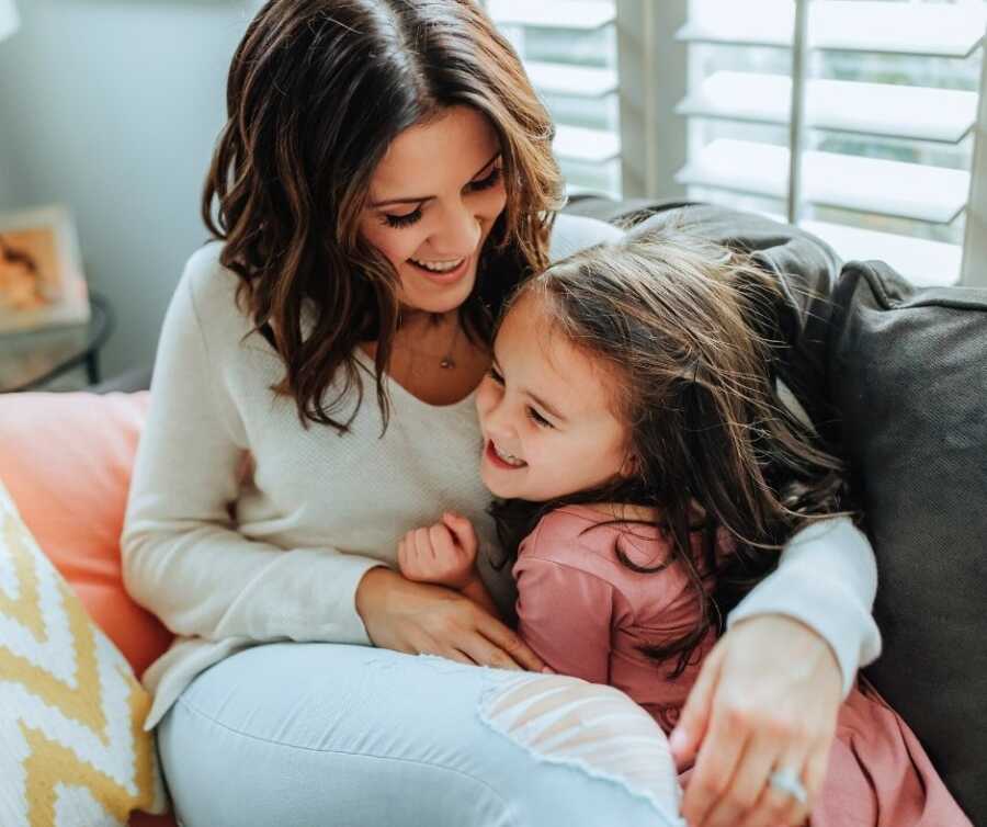 Mom and daughter sit together on the couch, smiling and laughing.