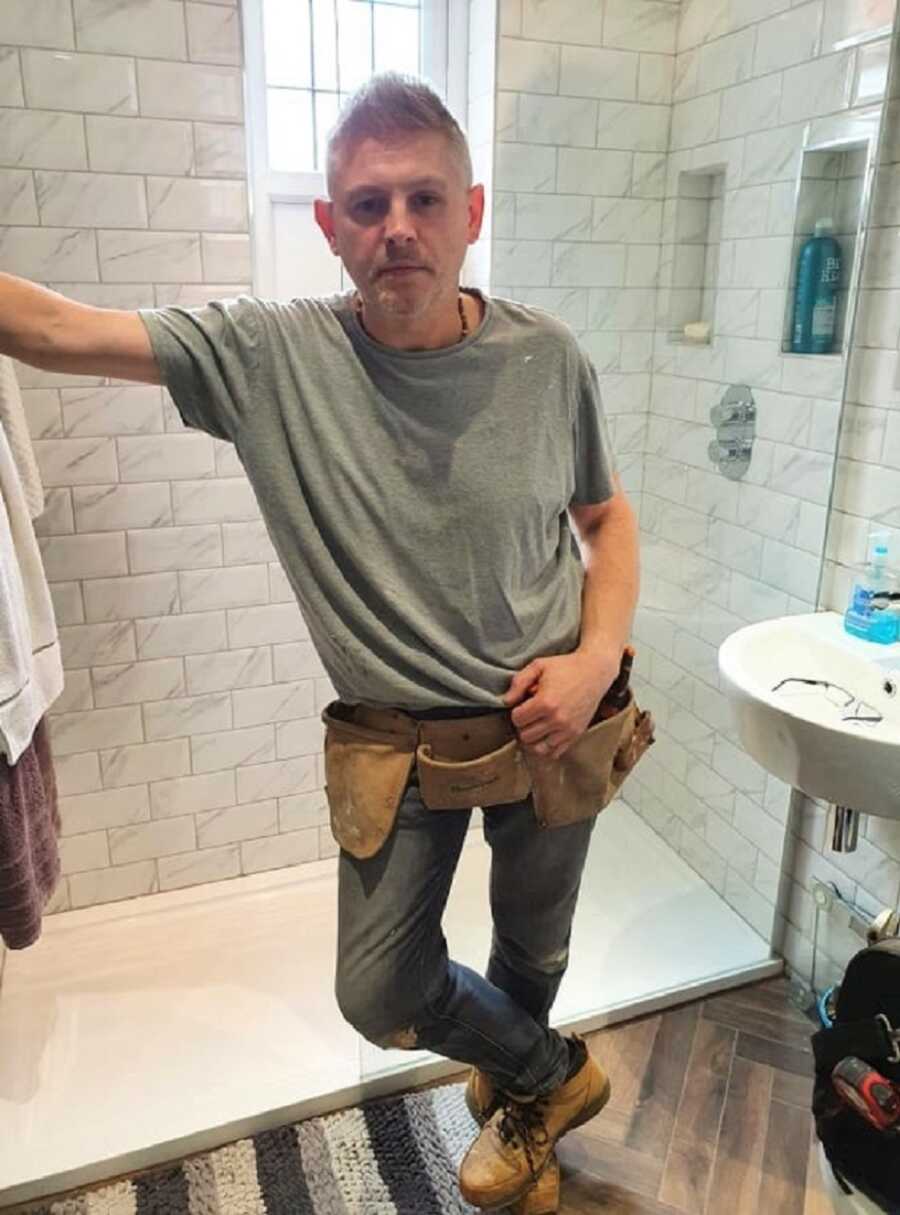 Male plumber poses in white tiled bathroom he has been working on. 