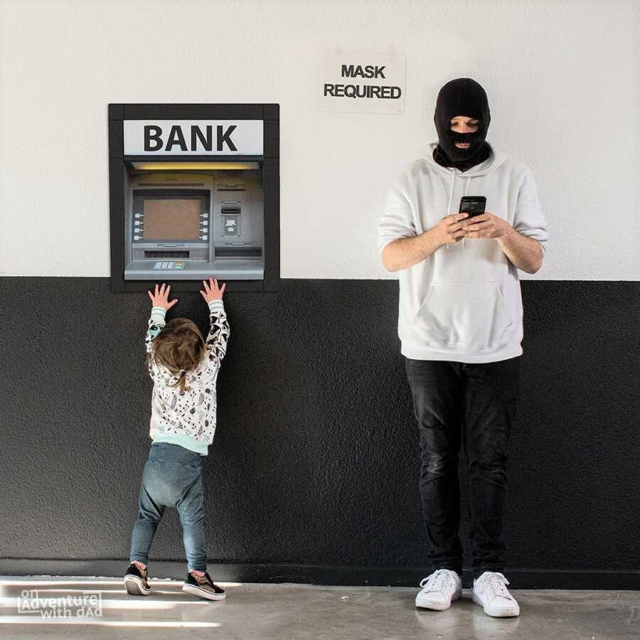 Dad wears black ski mask while toddler reaches for the bank's ATM.