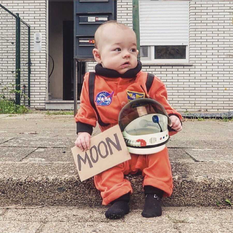 Small baby sits on the sidewalk curb in a space suit, trying to hitch hike to the moon.