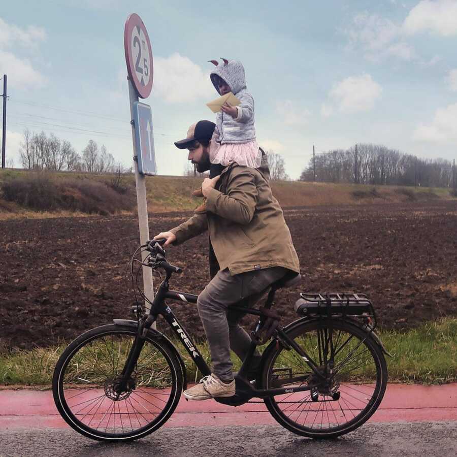 Dad rides bicycle with toddler balanced on his shoulders.