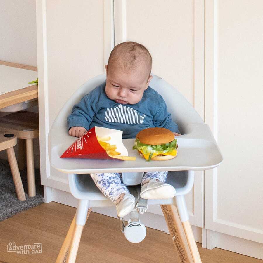 Dad jokingly feeds infant baby hamburger and fries. 