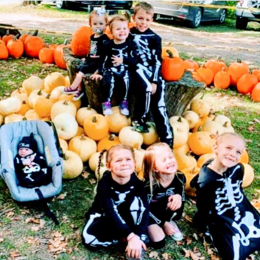 Two close-knit, blended families take a photo of their combined seven children, all in matching skeleton onesies for Halloween while sitting in a pumpkin patch