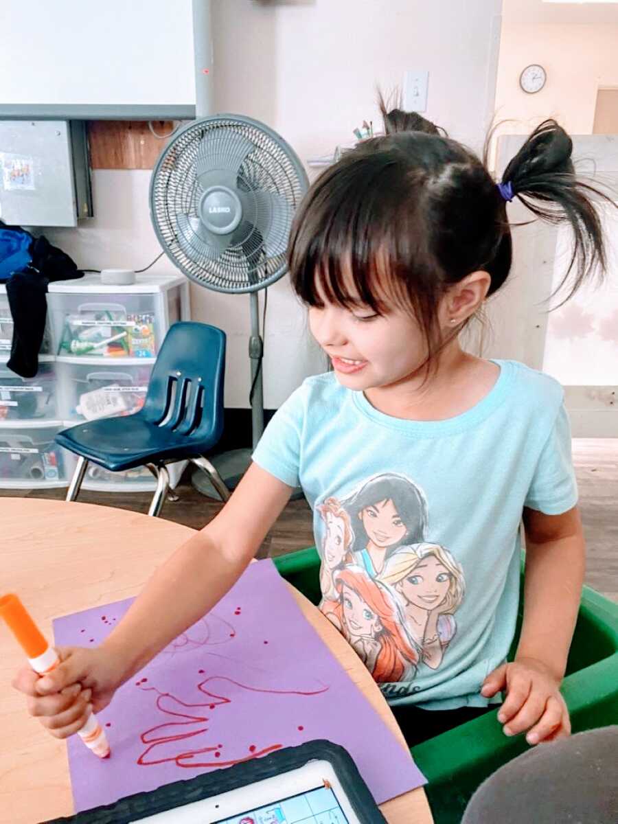 Little girl with autism sits at a table and colors on a purple piece of paper with an orange marker
