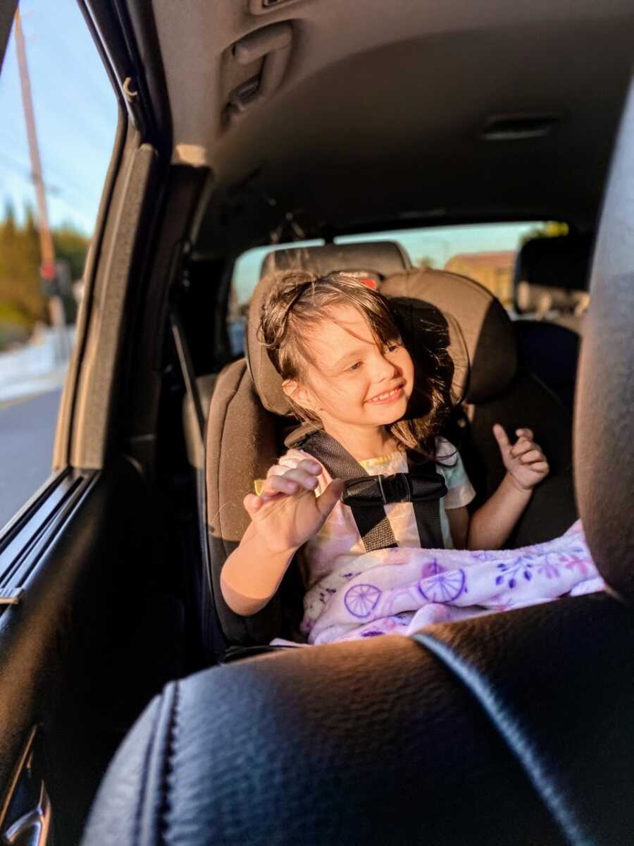 Little girl with autism smiles big while sitting in her car seat, the sun shining on her as wind blows through her hair