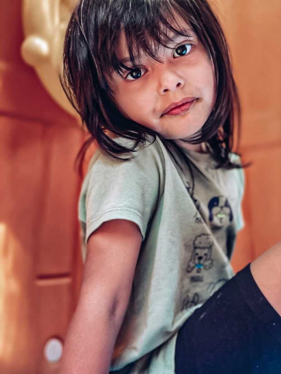 Little girl with brown hair and brown eyes looks intently into the camera while her mother takes a photo of her