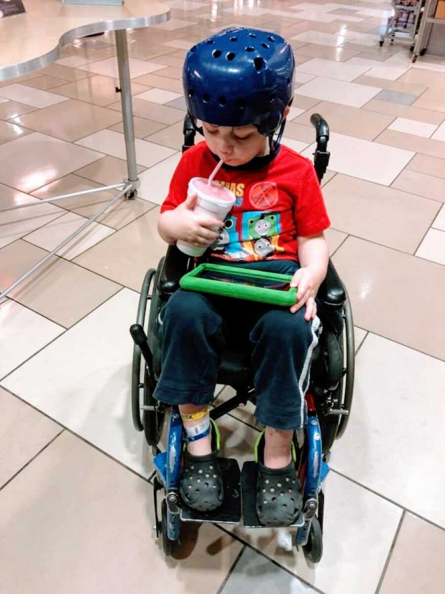 Little boy sits in a wheelchair and sips from a drink while wearing a blue helmet