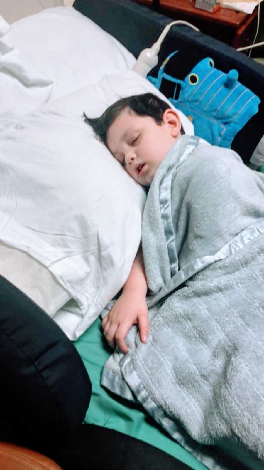 Little boy battling unknown seizures sleeps in a hospital bed with a Thomas the Train stuffed toy right next to him