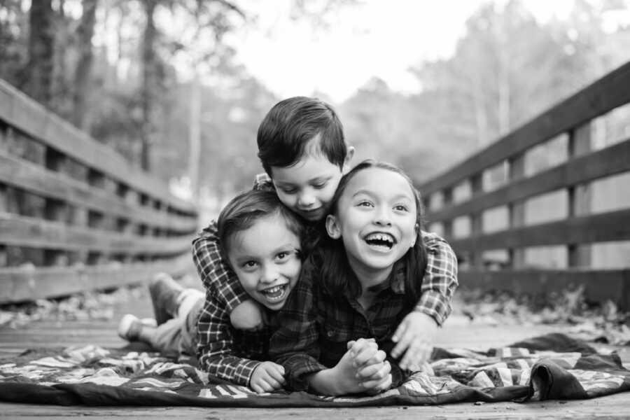 Three siblings take a photo together for a fall photoshoot while laying on a blanket on a wooden bridge