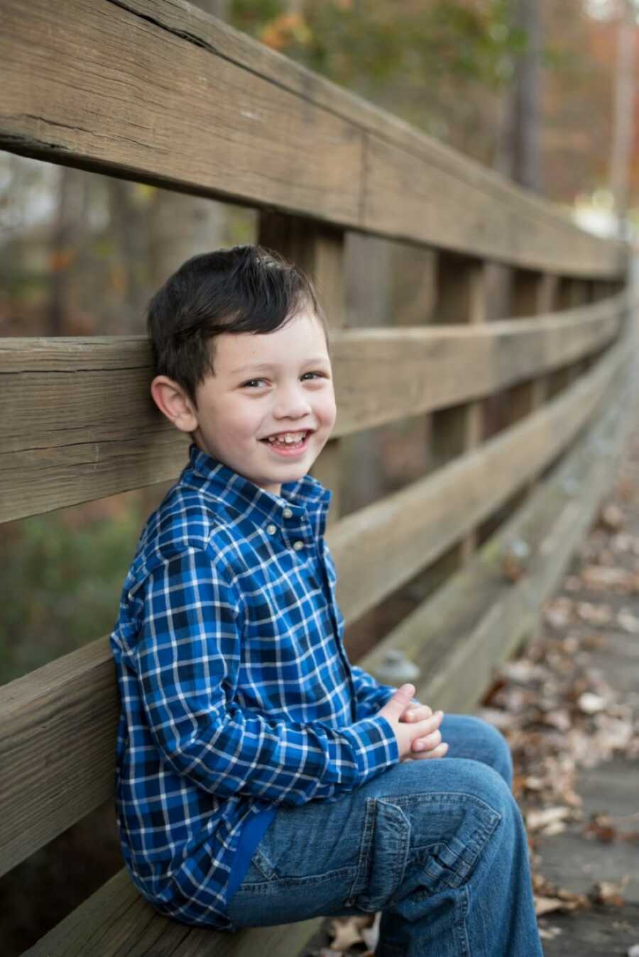 Little boy smiles big for a photo while wearing a blue plaid shirt and blue jeans, sitting down on a wooden bridge