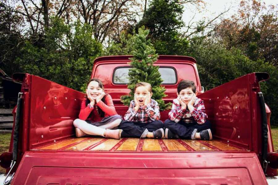 Mom shares Christmas photo of her three children sitting in the back of a red pick-up truck with a Christmas tree in it