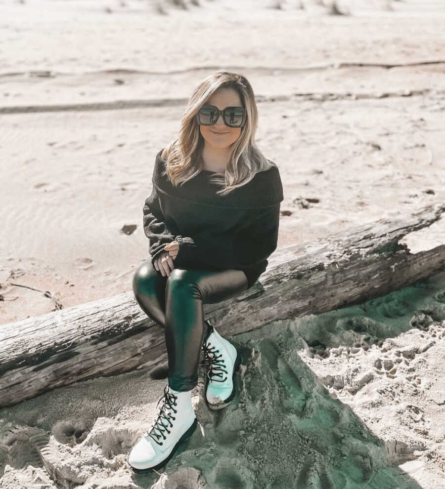 Woman on sobriety journey takes a photo at the beach in a black sweater, black pleather pants, and white combat boots