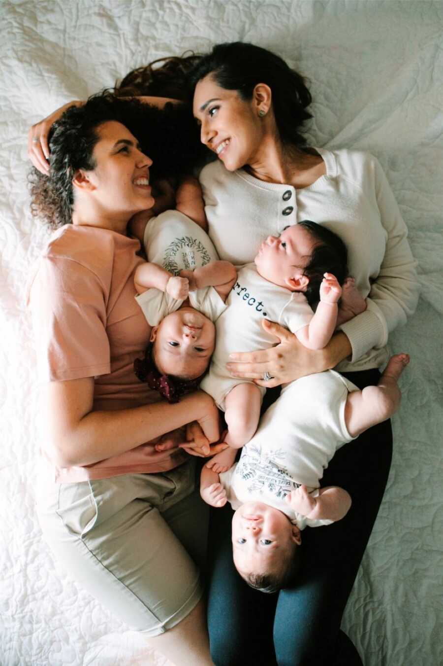 Two moms lay in bed and smile at each other while holding their triplet babies