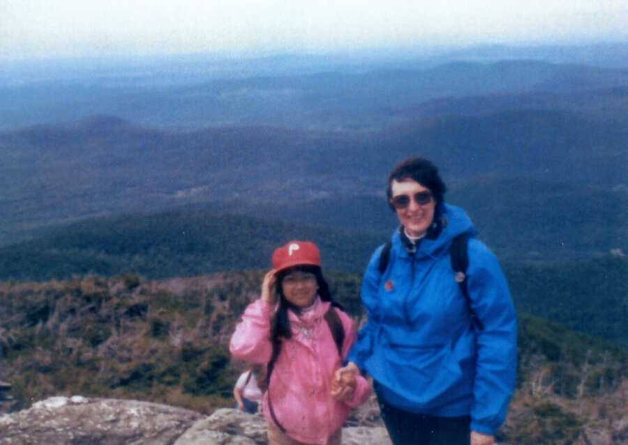 Young adoptee smiles for a photo with her adopted mom while on a family hike with mountains behind them