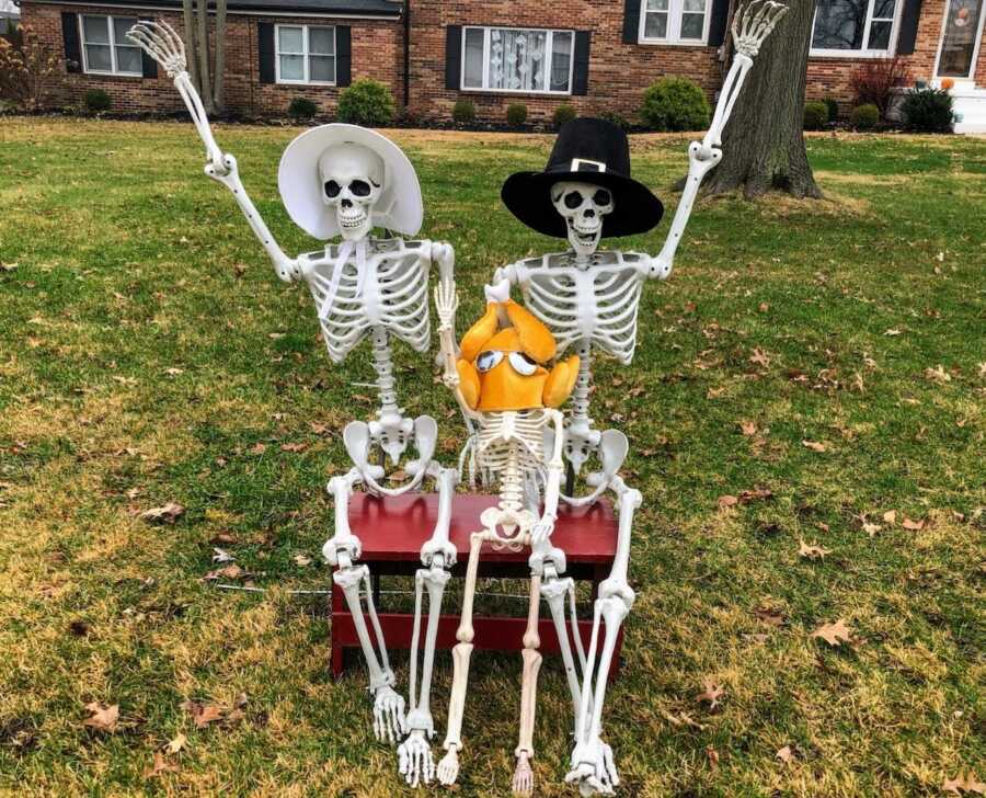 一对夫妇在他们的前院摆了三个骷髅装饰，戴着帽子庆祝感恩节