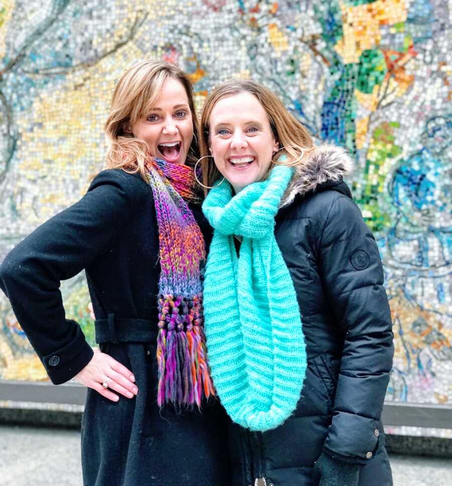 Two mom friends smile big for a photo in front a wall mural, both wearing black jackets and colorful scarves