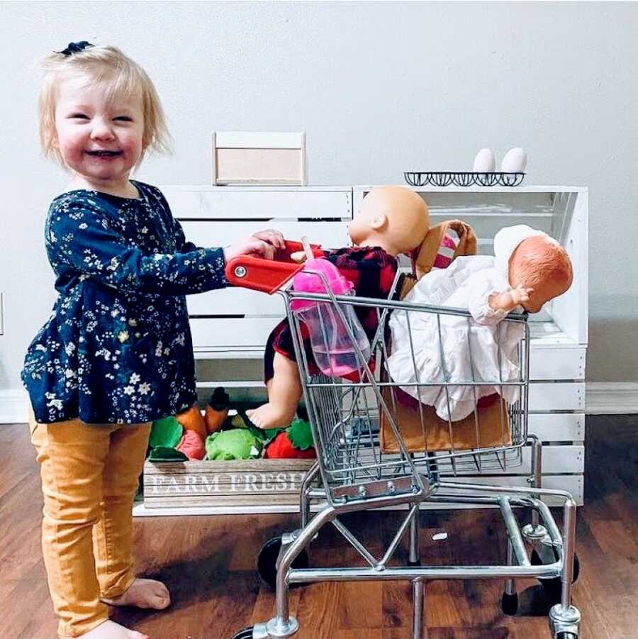 Little girl with Coffin-Siris Syndrome smiles big while playing pretend in a fake grocery store setup with a small buggy