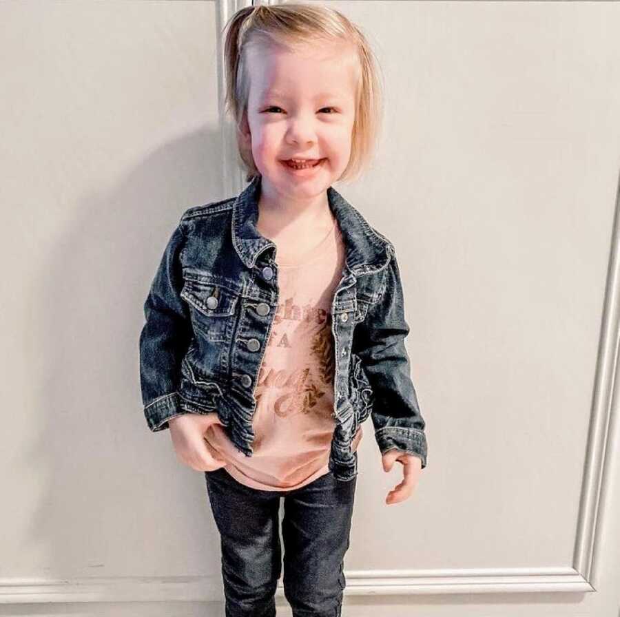 Little girl with Coffin-Siris Syndrome smiles big for a pictures while wearing a pink shirt under a denim jacket