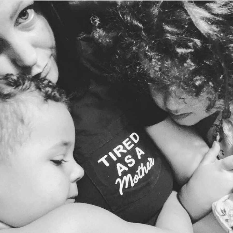 Single mom cuddles her two children while wearing a shirt that says "Tired as a mother"