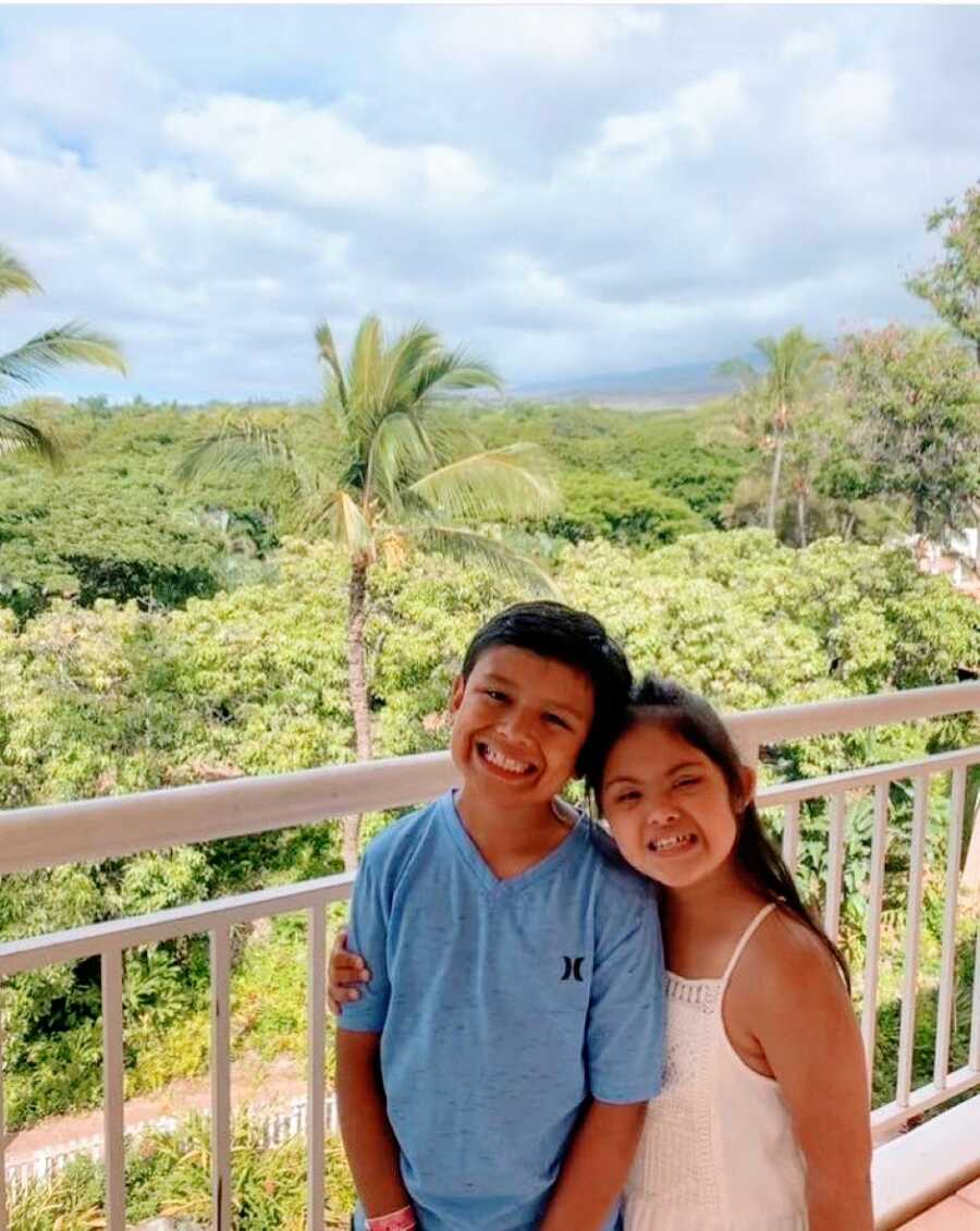 Brother and sister smile big and hug each other while standing on a balcony on a family vacation