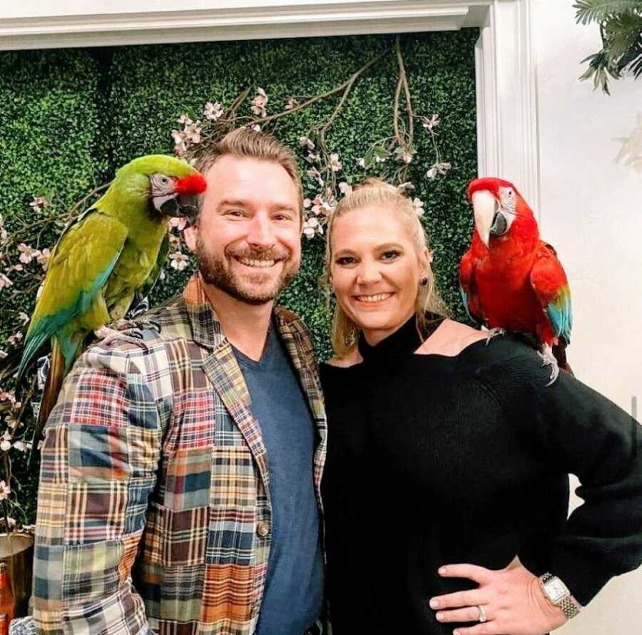 Husband and wife smile big while each have a parrot on their shoulder during a fun date night
