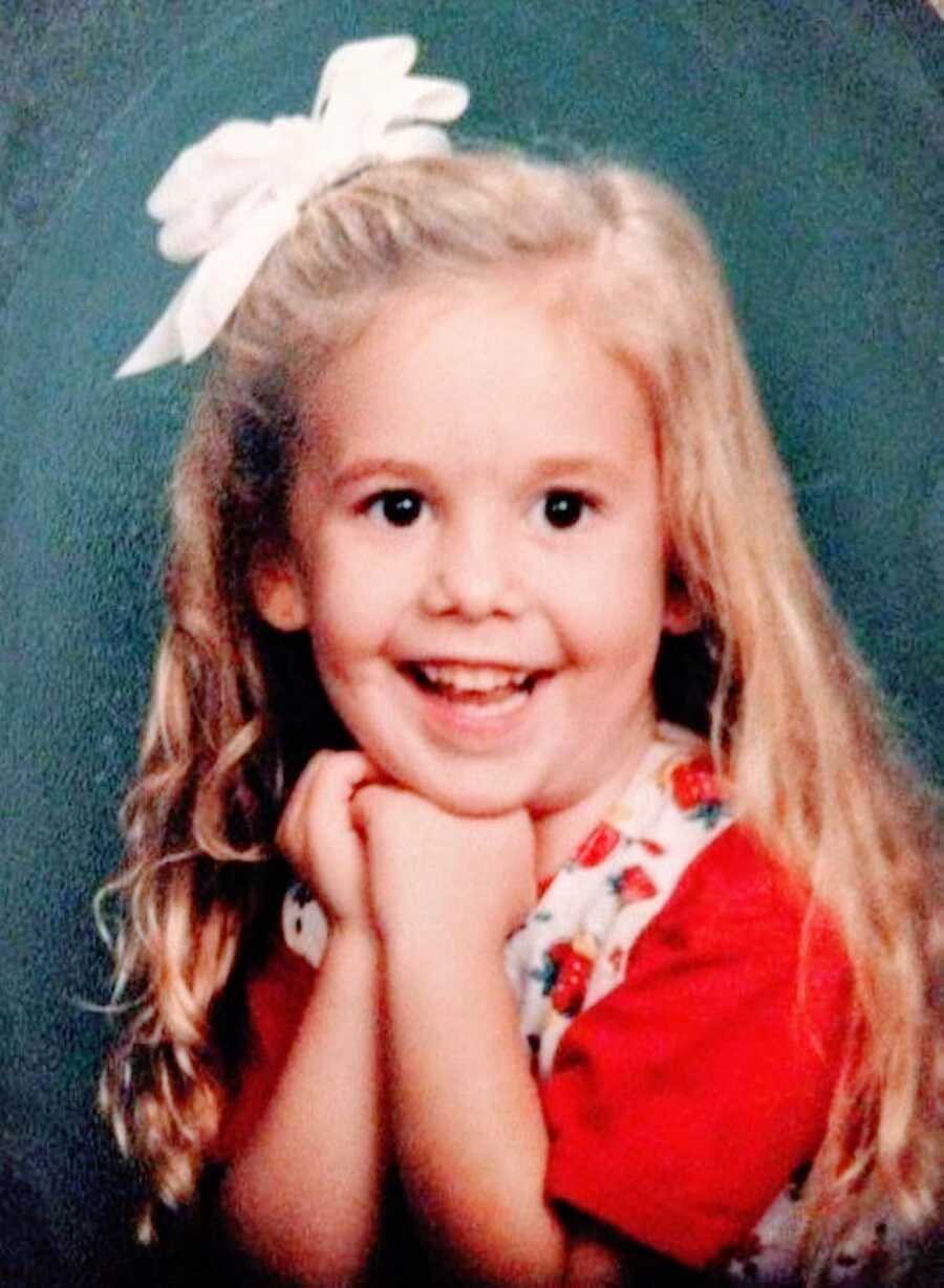 Little girl unknowingly battling OCD smiles for a school picture in a red dress with strawberries on it and a white bow in her hair
