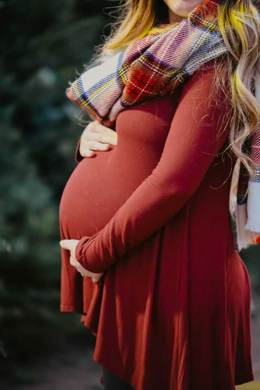 Very pregnant woman holds her baby bump while wearing a long-sleeved red dress and scarf