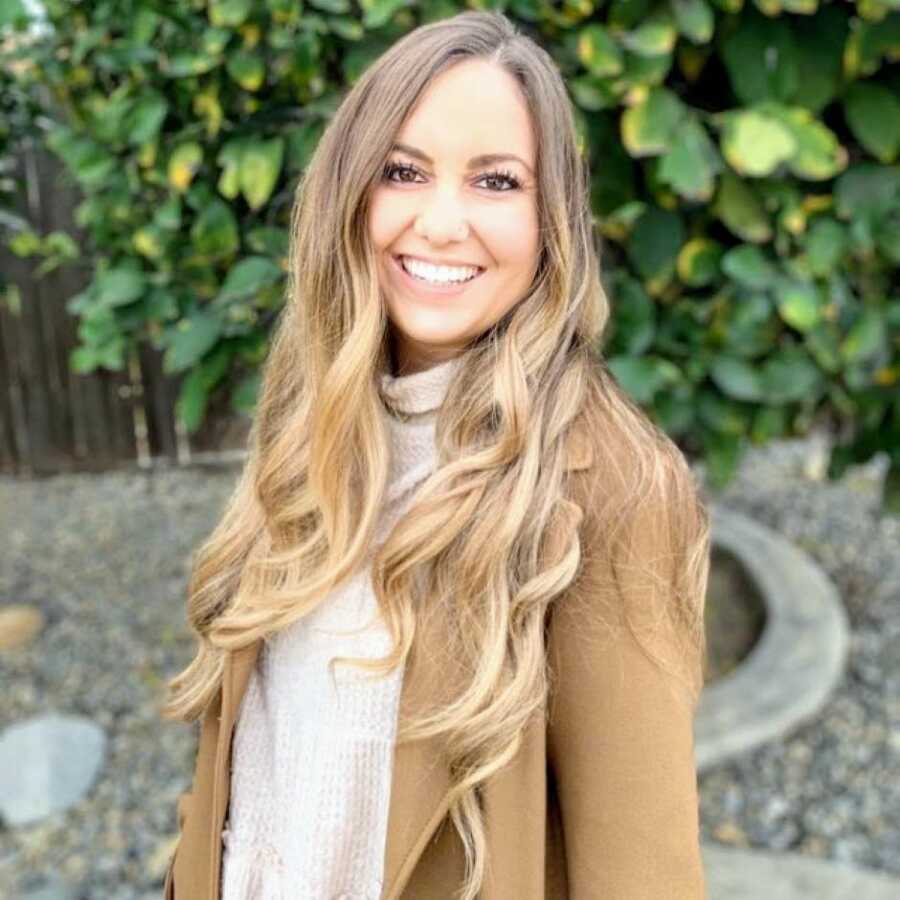 Mom and therapist battling OCD and anxiety smiles for a professional headshot in a tan sweater and brown jacket