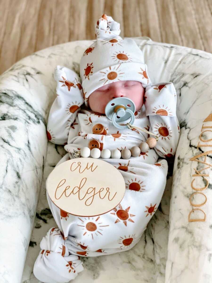 Newborn baby boy sucks a pacifier while wearing a matching onesie and beanie with suns on it