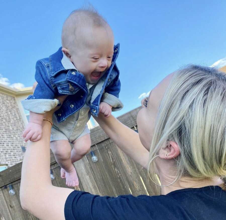 Mom plays airplane in her backyard with her young, happy and smiling son with Down syndrome