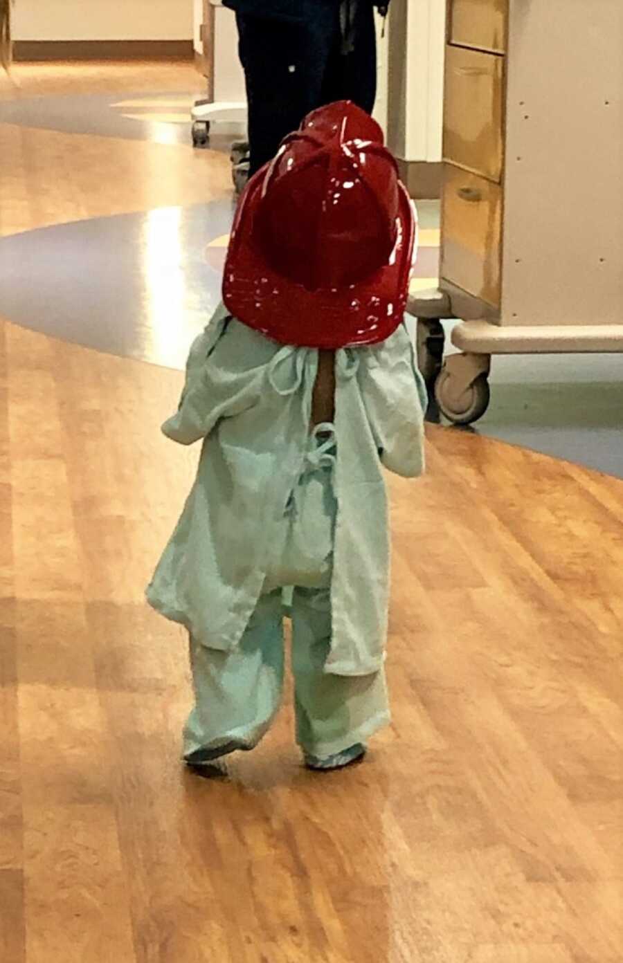 A young patient in hospital clothing walks away wearing a firefighter hat.