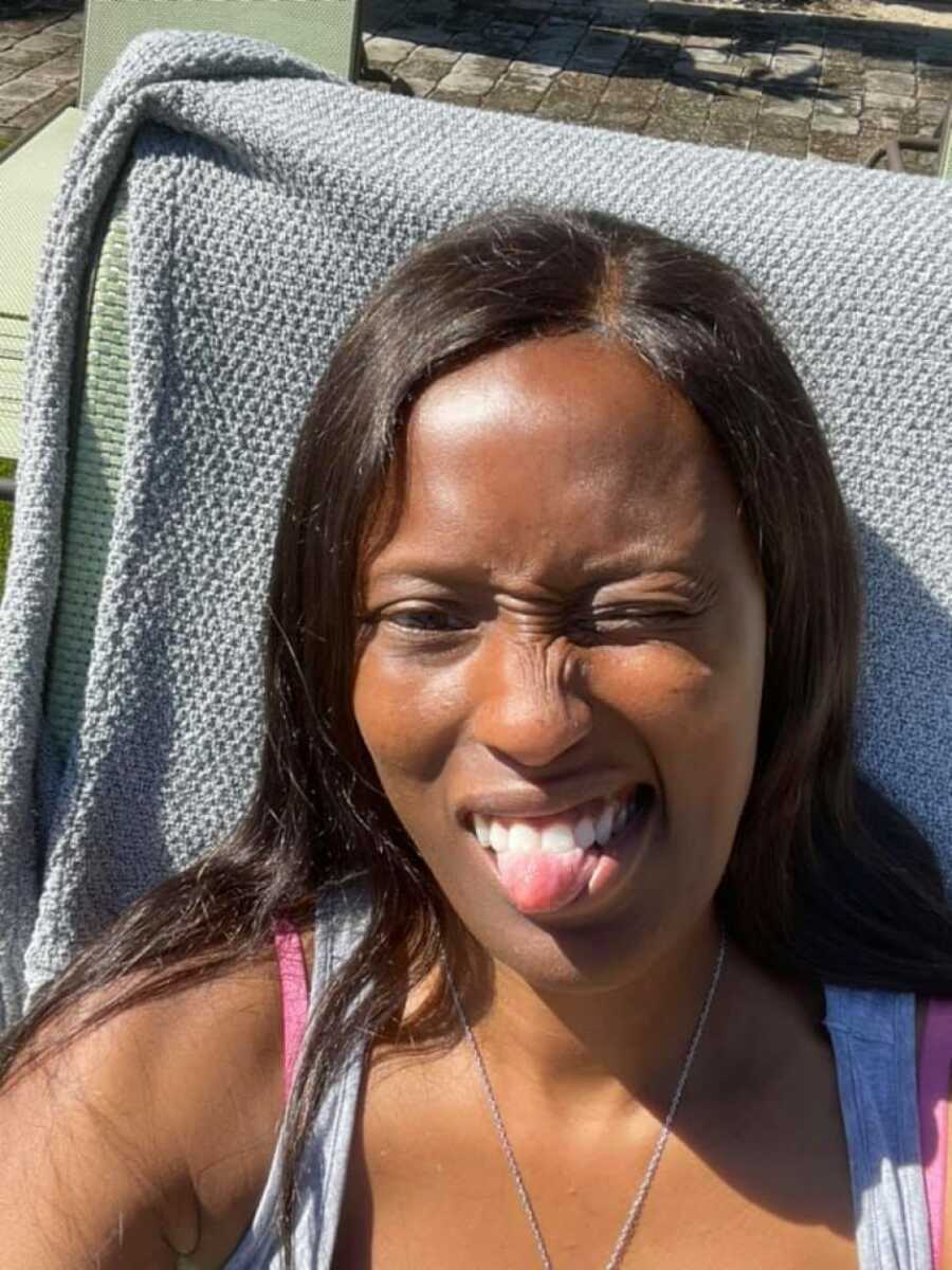 woman with long dark hair is resting against a grey chair with her tongue sticking out