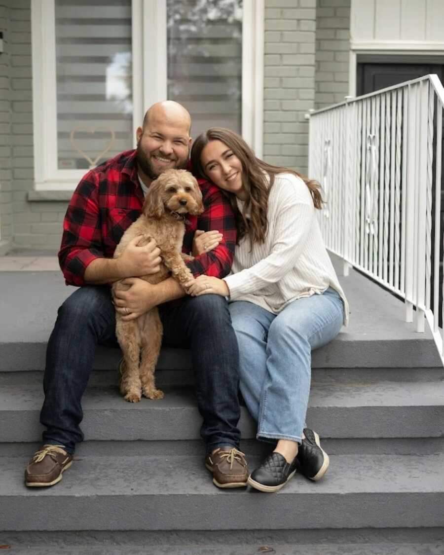 Couple sitting on their grey steps in front of their house while holding their adorable dog