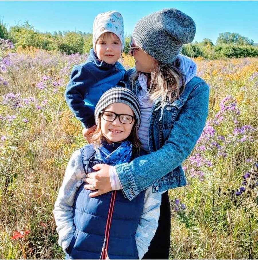 Mom stands with her two earth-side sons in a field with wild flowers swaying in the wind behind them
