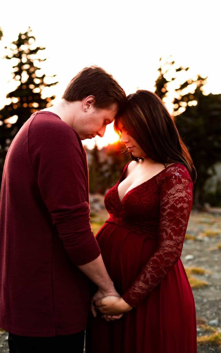 Teen couple take maternity photos together in matching maroon outfits while the sun sets in the trees behind them