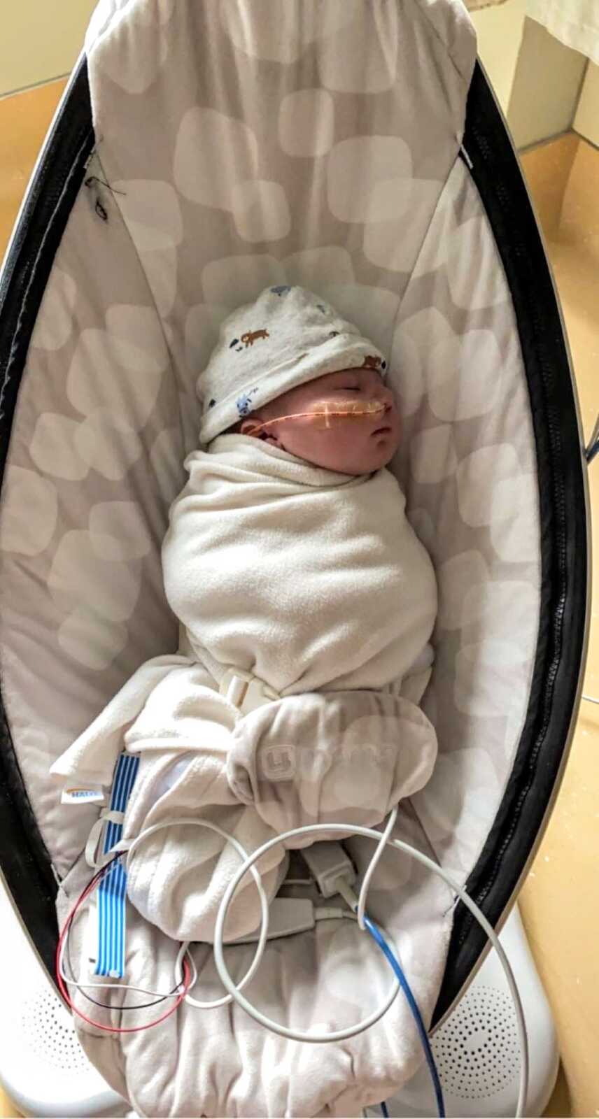Newborn boy swaddled in a white blanket sleeps in a baby swing with a feeding tube